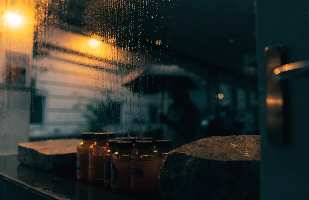 Rain-streaked pharmacy window at night with warm street light spilling across the glass